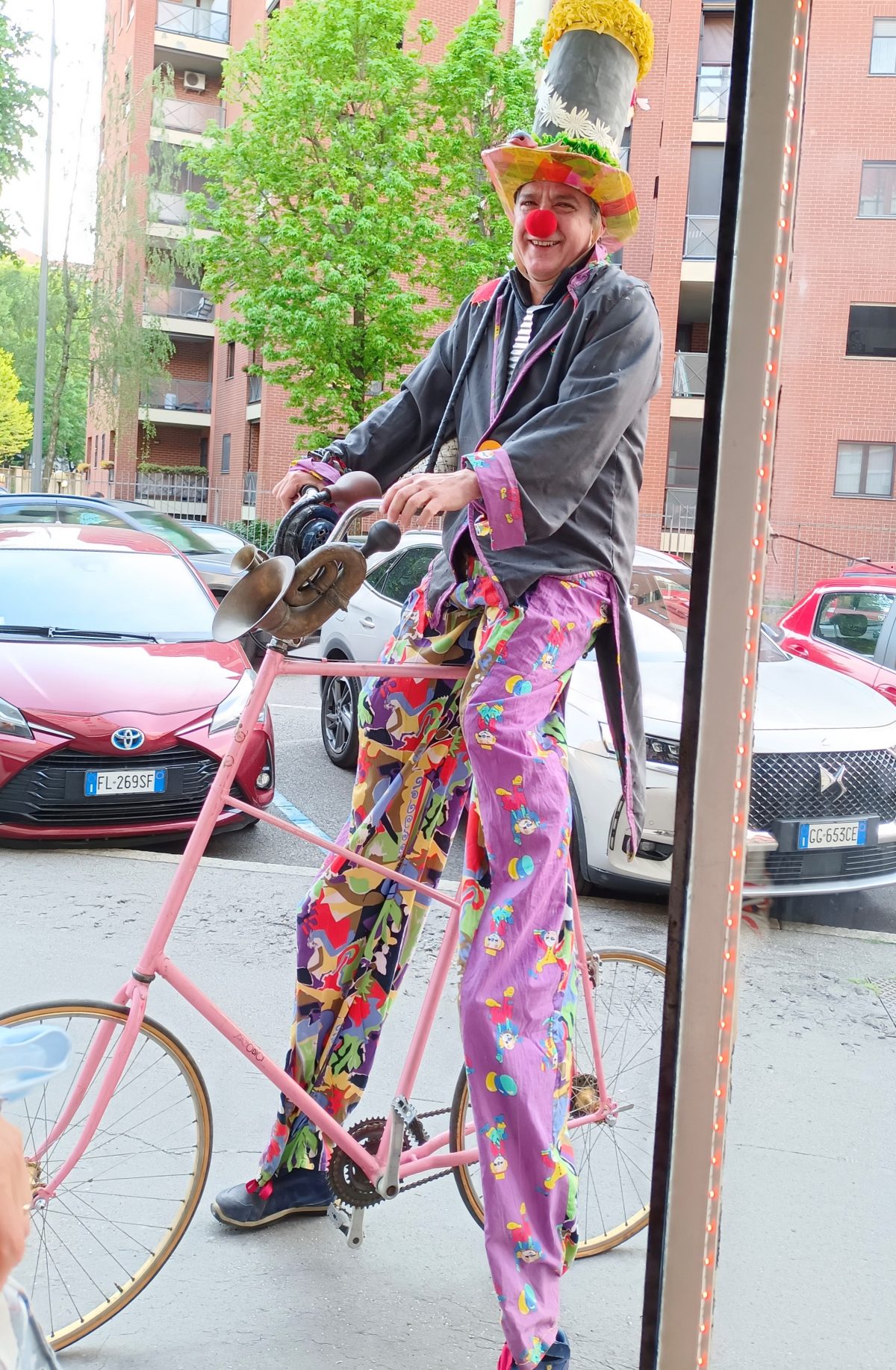 A stilt-clown in patterned trousers on a pink bicycle, performing outside the storefront